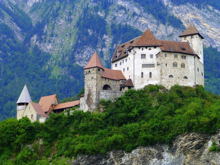 Burg mit roten Ziegeldächern auf einem bewaldeten Felsen vor einer Bergwand